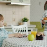 Caring grandmother is bringing plate with fresh croissants feeding granddaughter at home in kitchen, child is clapping hands and smelling pastry